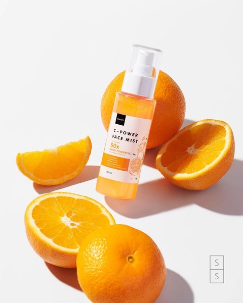 A high-quality product shot of a face mist spray bottle surrounded by halved and sliced fresh oranges, emphasizing natural ingredients.